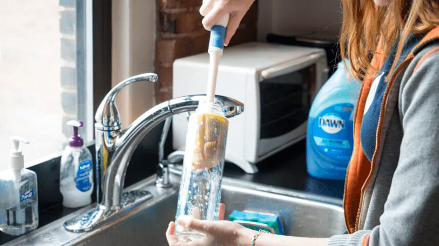 person cleaning clear water bottle with brush in a stainless steel sink